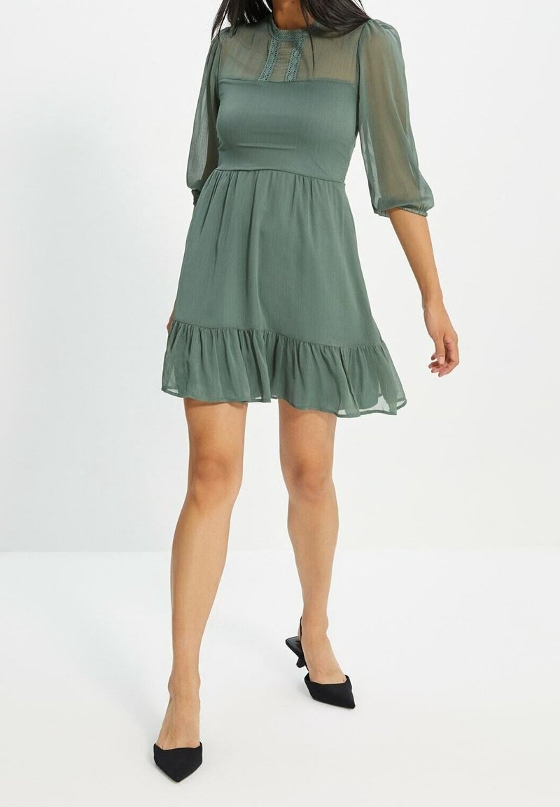 Femme portant une courte robe verte avec des manches bouffantes transparentes et des talons noirs pointus, debout sur un fond blanc.