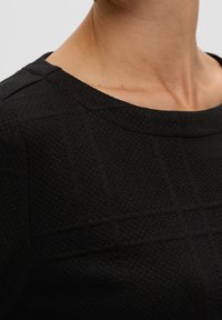 Tissu noir texturé avec un motif carré, présentant un encolure bateau et des bords lisses autour des épaules, montrant des détails subtils.