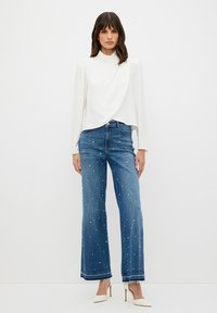 Blusa bianca a maniche lunghe con scollo alto, abbinata a jeans a gamba larga blu con decorazioni in perle e orlo sfrangiato. Tacchi bianchi.