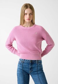 Pink ribbed sweater with a round neck and long sleeves, featuring a small logo patch. Paired with high-waisted blue denim jeans.