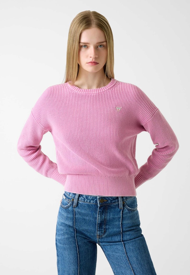 Pink ribbed sweater with a round neck and long sleeves, featuring a small logo patch. Paired with high-waisted blue denim jeans.