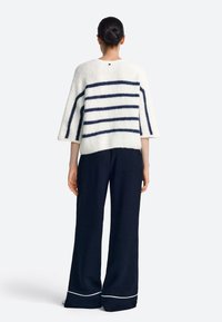 Fehér csíkos pulóver tengerészkék vonalakkal, texturált anyagból, kerek nyakkal és háromnegyedes ujjakkal, laza fekete nadrággal, fehér szegéllyel párosítva.