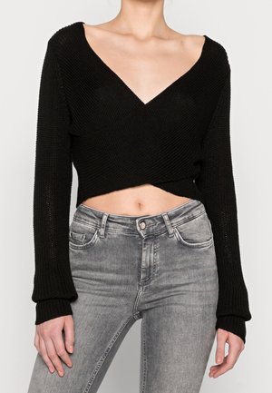 Femme portant un pull court à manches longues noir avec un col en V profond et un jean gris taille haute.