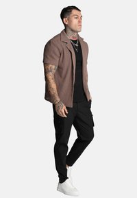 Brun kortärmad, texturerad skjorta med knappar över en svart ribbad tröja, kombinerad med svarta cargo-byxor och vita sneakers.