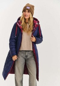 Pikowany długi płaszcz w ciemnym granacie z kwiatową podszewką, zapinany na zamek błyskawiczny, w połączeniu z beżowym swetrem i szarymi dżinsami. Brązowa dziana czapka.