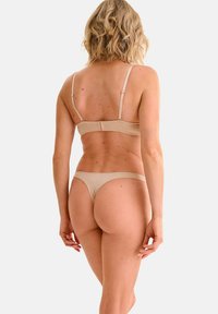 Donna con capelli biondi corti che indossa un reggiseno beige e un perizoma, in piedi contro uno sfondo bianco semplice, rivolta di spalle verso lo spettatore.