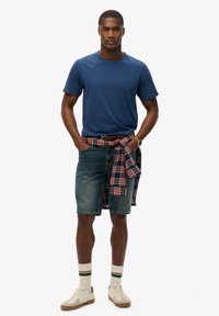 Homme portant un t-shirt bleu marine, un short en jean, des baskets blanches avec des chaussettes rayées, et une chemise à carreaux nouée autour de la taille.