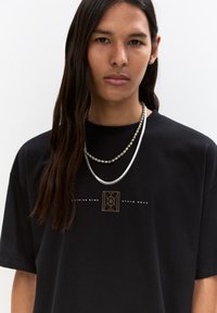 T-shirt in cotone nero con una grafica centrale marrone, collo rotondo, maniche corte. Due collane sovrapposte, una in argento e una con perline.