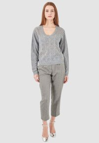 Donna con lunghi capelli rossi che indossa un maglione grigio a V in tessuto testurizzato, pantaloni a scacchi grigi alla caviglia e tacchi con cinturino argentato, in piedi contro uno sfondo bianco.