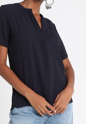 Vrouw draagt een marineblauwe kortarmblouse met kleine rode stippen en lichtblauwe spijkerbroek, handen gevouwen bij de taille.