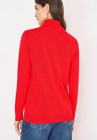 Pull-over rouge à col montant, manches longues, et texture lisse. L'ourlet est côtelé, il est associé à un jean en denim bleu.