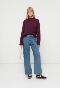 Maglione a maglia bordeaux, con colletto alto, jeans larghi blu, sneakers nere con dettagli bianchi, mentre tiene una piccola borsa bianca.