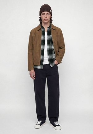 Jeune homme portant une veste marron, une chemise à carreaux noirs et blancs, un pantalon noir, un bonnet noir et des baskets noires, debout devant un fond blanc.