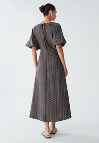 Brun maxikjole med hvide prikker, flagrende ærmer, nøglehul i ryggen og en afslappet pasform, lavet af letvægtsstof. Flade sandaler er iført.