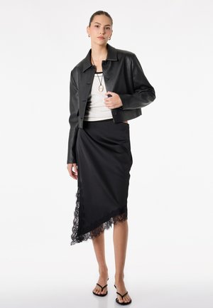 Donna che indossa una giacca di pelle nera corta, top bianco, gonna nera asimmetrica con rifinitura in pizzo, sandali neri e collana con ciondolo su sfondo bianco.