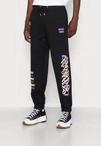 Person som bär svarta sweatpants med gul och lila grafiktext och svarta högskoor med tjocka vita sulor.