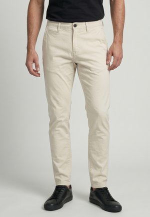 Homme portant un chino beige slim fit et des baskets noires, debout devant un fond clair uni.