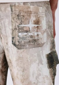 Vernietigde denim in beige met een camouflagepatroon. Bevat een patchzak met een wit logo en tekst met de vermelding "Project X Paris."