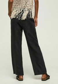 Czarne lniane spodnie o szerokich nogawkach z gładką teksturą, z wykończonymi krawędziami i noszone w połączeniu z czarnymi sandałami. Widok z tyłu ukazuje luźny krój.