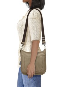 Helles Leder-Umhängetasche mit weicher Textur, zwei Vorderfächer und einem braunen verstellbaren Gurt, als Crossbody-Tasche an der Seite der Frau getragen.