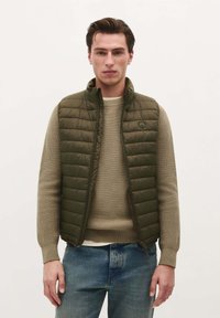 Vestă pufos de culoare verde măslinie, cu un model matlasat, purtată cu un pulover bej cu model canalat și blugi dungati albaștri. Textură fină, guler înalt și fermoar.