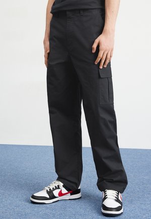 Hombre con pantalones cargo negros y zapatillas Nike blancas, negras y rojas, de pie sobre una alfombra azul contra una pared blanca.