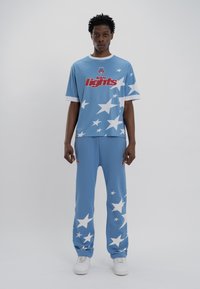 Ανοιχτό μπλε σετ αθλητικών ρούχων που περιλαμβάνει ένα μπλουζάκι κοντομάνικο και ταιριαστά παντελόνια, και τα δύο με μεγάλα λευκά σχέδια αστεριών και εκτυπωμένο κόκκινο κείμενο.