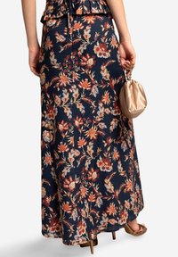 Maxi-Rock aus dunkelblauem Stoff mit roten und orangefarbenen Blumen. Verfügt über ein fließendes Design und einen Schlitz, kombiniert mit einer beigefarbenen Clutch.
