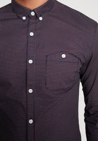 Camicia bordeaux scuro con bottoni, con un piccolo motivo geometrico, bottoni bianchi e taschino sul petto. Tessuto di cotone con una trama liscia.