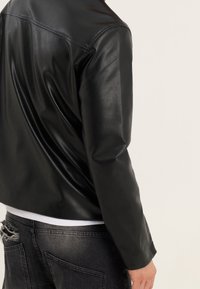 Giacca di pelle nera con una texture liscia, caratterizzata da un taglio classico e cuciture minime. Indossata sopra una camicia bianca e jeans scuri strappati.