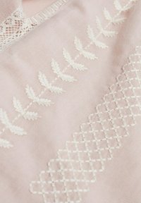 Tissu rose présentant une broderie blanche complexe de feuilles et un motif géométrique, avec une délicate bordure en dentelle le long de l'encolure.