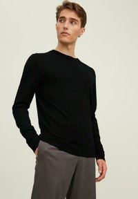 Maglione nero a maglia con maniche lunghe e scollatura a girocollo, caratterizzato da una trama liscia e una silhouette aderente. Indossato con pantaloni grigi.
