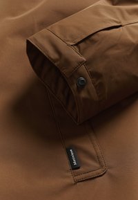 Podrobnost rjave vodoodporne jakne, s podvrnjeno manšeto s črnim pritisnim gumbom in etiketo blagovne znamke, izdelana iz gladkega materiala z poudarki šivanja.