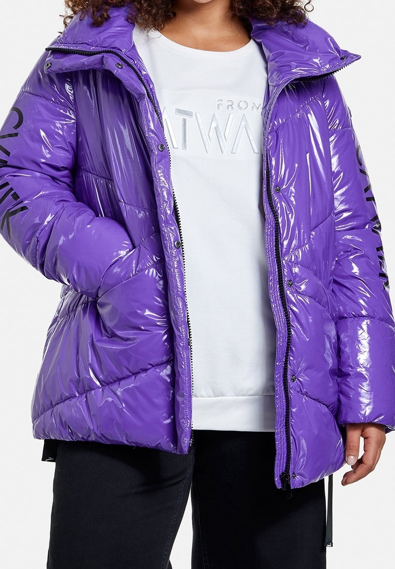 Fialová, lesklá péřová bunda se zipem, kapucí a černým potiskem na rukávech. Má prošívaný design pro izolaci.