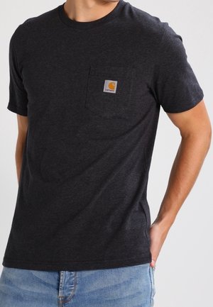 Homme portant un t-shirt à manches courtes Carhartt gris foncé avec une poche poitrine et un jean bleu clair, debout avec une main dans sa poche.