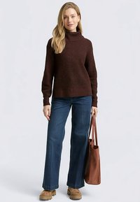 Brauner, gerippter Rollkragenpullover mit langen Ärmeln, kombiniert mit blauen Schlagjeans und beigen Plateau-Stiefeln. Modell hält eine braune Tragetasche.