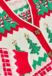 Gestrickter Urlaubspullover mit roten Abschlüssen, grünen Weihnachtsbaum-Mustern, goldenen Paillettensternen und braunen Knöpfen auf weißem und rotem Hintergrund.