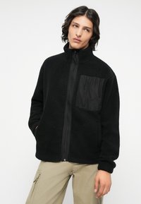Junge Person trägt eine schwarze Fleecejacke mit Brusttasche und beige Cargo-Hosen, steht mit Händen in den Taschen vor weißem Hintergrund.