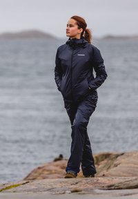 Navy waterproof jacket with hood and zipper pockets, paired with matching pants, worn while walking on rocky shoreline near water.
