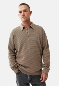 Beige pitkähihaine polo-paita, jossa on teksturoitu kuvio, kolme nappia kauluksessa ja resorit, yhdistettynä tummiin, väljiin farkkuihin.