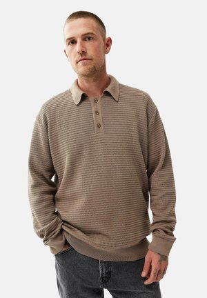 Beige langærmet polo shirt med tekstureret mønster, tre knapper ved kraven og ribbede ærmer, parret med mørke, løstsiddende jeans.