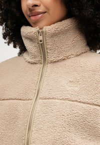 Beige fleece jacket featuring a high collar, full zipper, and textured fabric. Embroidered logo detail near the chest.