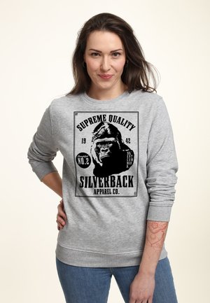 Kobieta o ciemnych włosach nosząca szarą bluzę z czarną grafiką goryla i napisem "Supreme Quality Silverback Apparel Co."