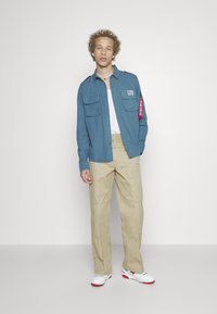 Modrá košeľa s dlhým rukávom a dvoma prednými vreckami, béžové nohavice a biele tenisky s červenými akcentmi. Obsahuje štítok na rukáve.