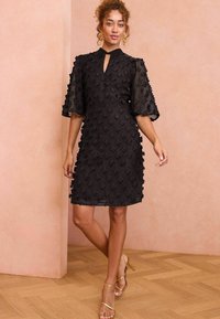 Rochie neagră cu un model floral texturat, manșete scurte din plasă și decolteu cu deschidere în formă de cheie. Asortată cu sandale cu toc metalice cu bretele.