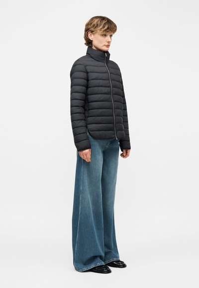 Άτομο που φοράει μαύρο μπουφάν με καπιτονέ ύφασμα και μπλε τζιν παντελόνι με φαρδύ πόδι, στέκεται πλευρικά σε λευκό φόντο.
