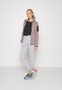 Rød og grå mønstret zip-up jakke med sidelommer, sort t-shirt, grå sweatpants og blå-orange sportsko.