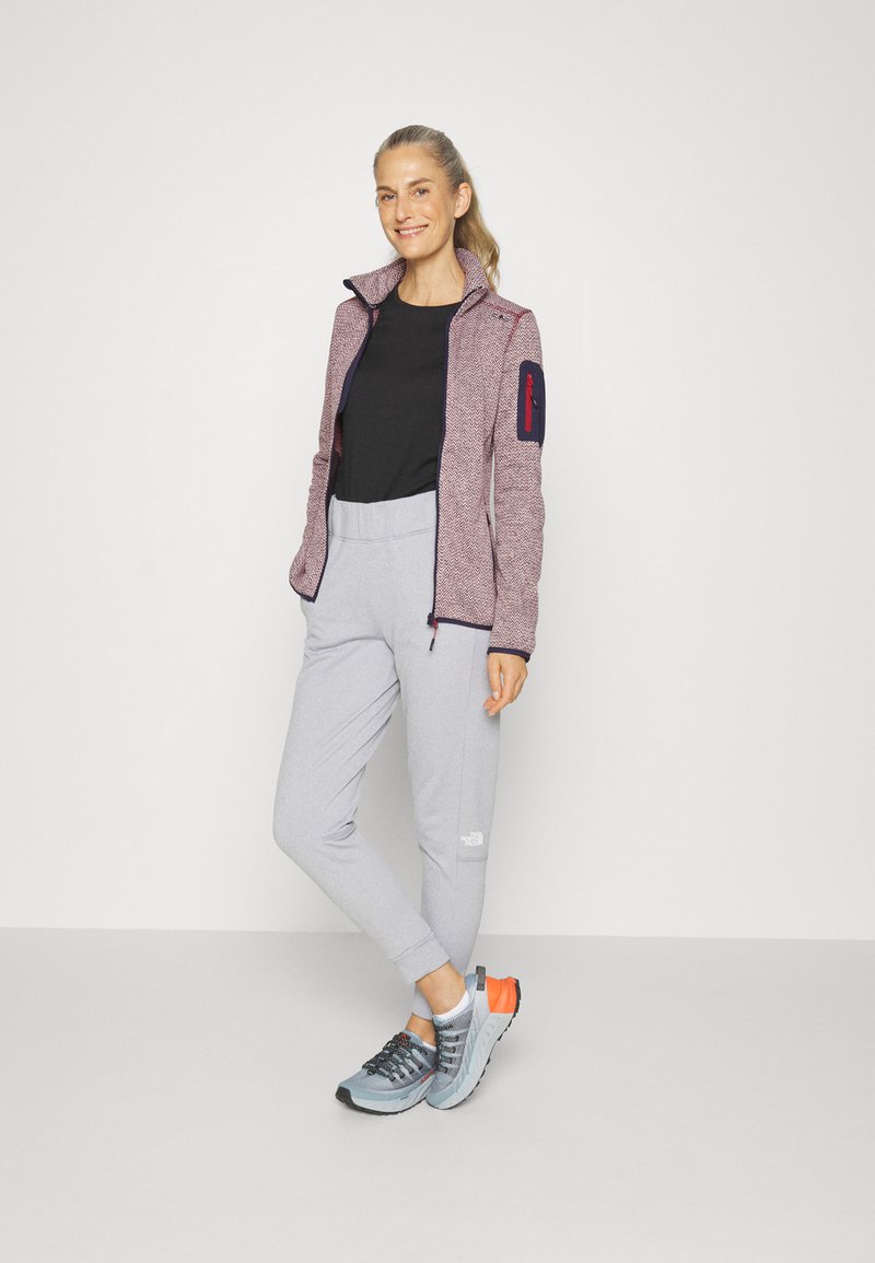 Giacca zip-up rossa e grigia con motivo e tasche laterali, t-shirt nera, pantaloni della tuta grigi e scarpe da ginnastica blu-arancioni.