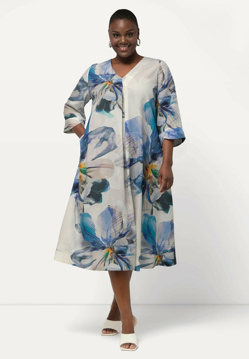 Vestido de estampado floral con mangas tres cuartos, escote en forma de V y bolsillos. Tejido ligero con patrones florales en azul, verde y amarillo sobre un fondo blanco.