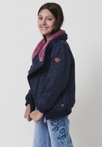 Giovane donna sorridente che indossa una giacca patrimoniale blu navy con fodera rosa e jeans azzurri chiari con ricami floreali, in piedi di lato.